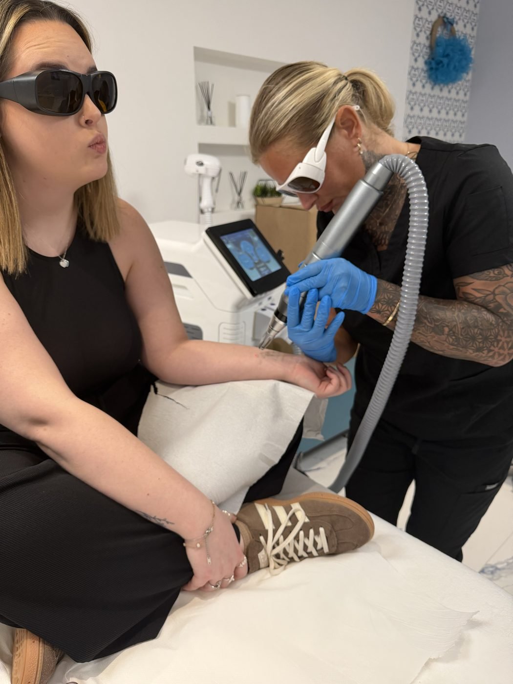 Supervised laser tattoo removal training, a practitioner treating a forearm tattoo while the learner observes, both wearing laser safety eyewear